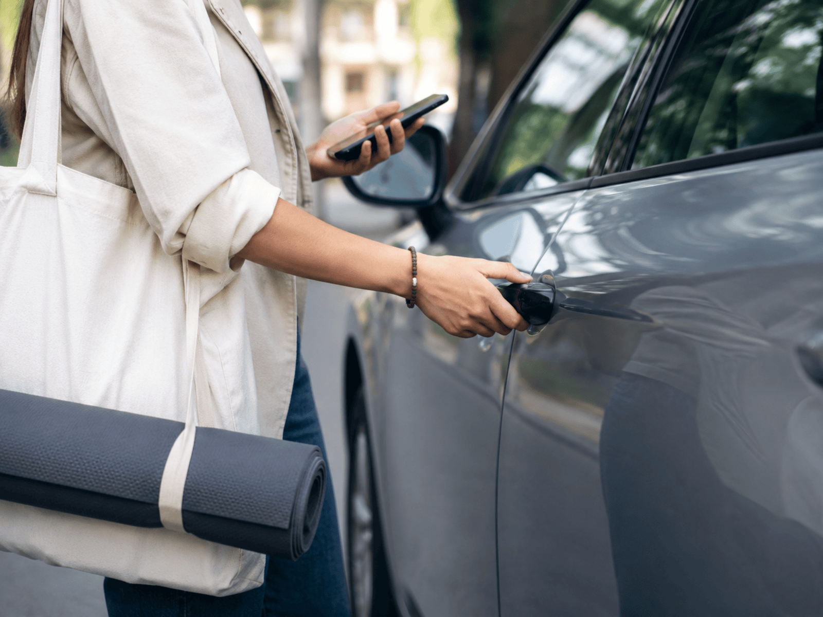 Una donna con una borsa e un tappetino da yoga a tracolla tiene il cellulare nella mano sinistra e con la destra apre la portiera di un’auto grigia.