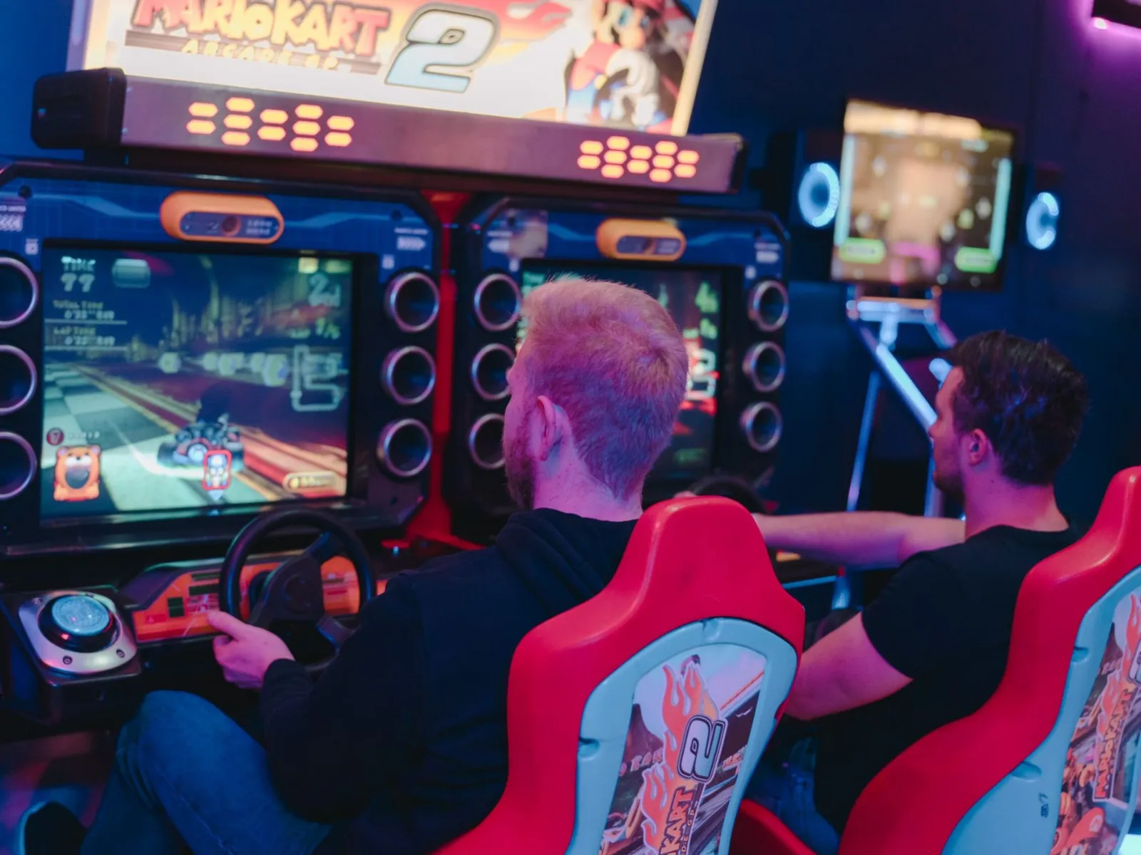 İki adam kırmızı atari yarış koltuklarına oturmuş, bir atari salonunda "Mario Kart Arcade GP 2" oynuyorlar.