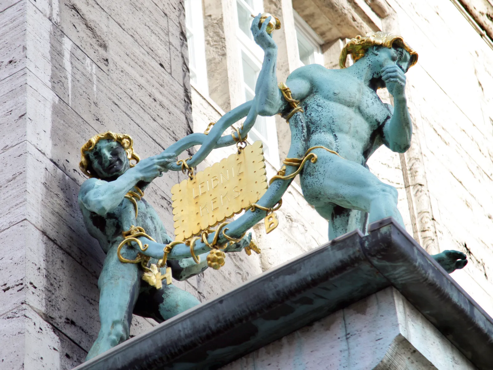 Dos figuras de bronce portan un pretzel de bronce. En este pretzel cuelga una galleta dorada con la inscripción "Galleta Leibniz".