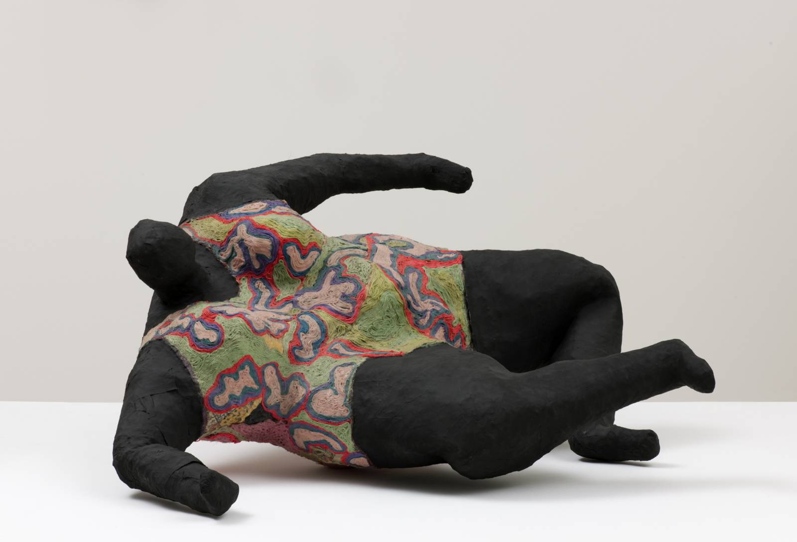 Zu sehen ist eine Nana-Puppe von Niki de Saint Phalle. Sie hat einen schwarzen Körper, liegt seitlich auf einer weißen Platte und trägt einen Badeanzug ähnliches Kleidungsstück, das mit einem organischen Muster überzogen ist.