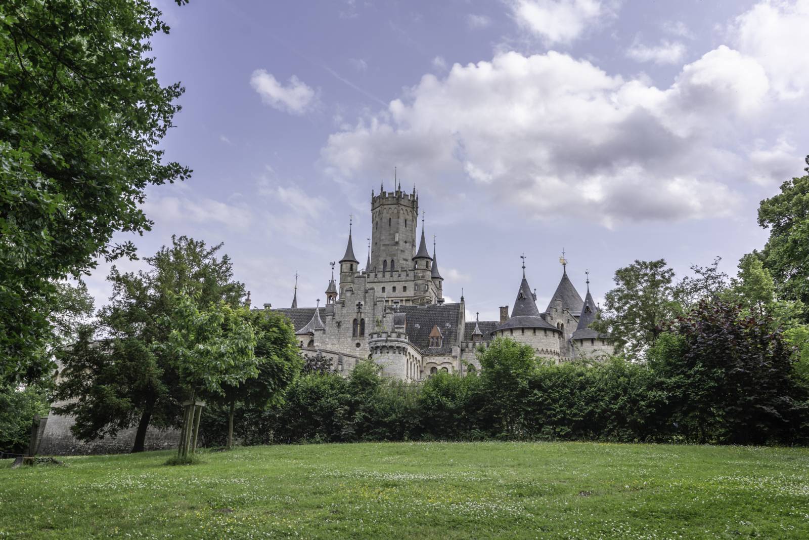 Blick auf das märchenhafte Schloss Marienburg hinter Bäumen und einer grünen Wiese an einem leicht bewölkten Tag