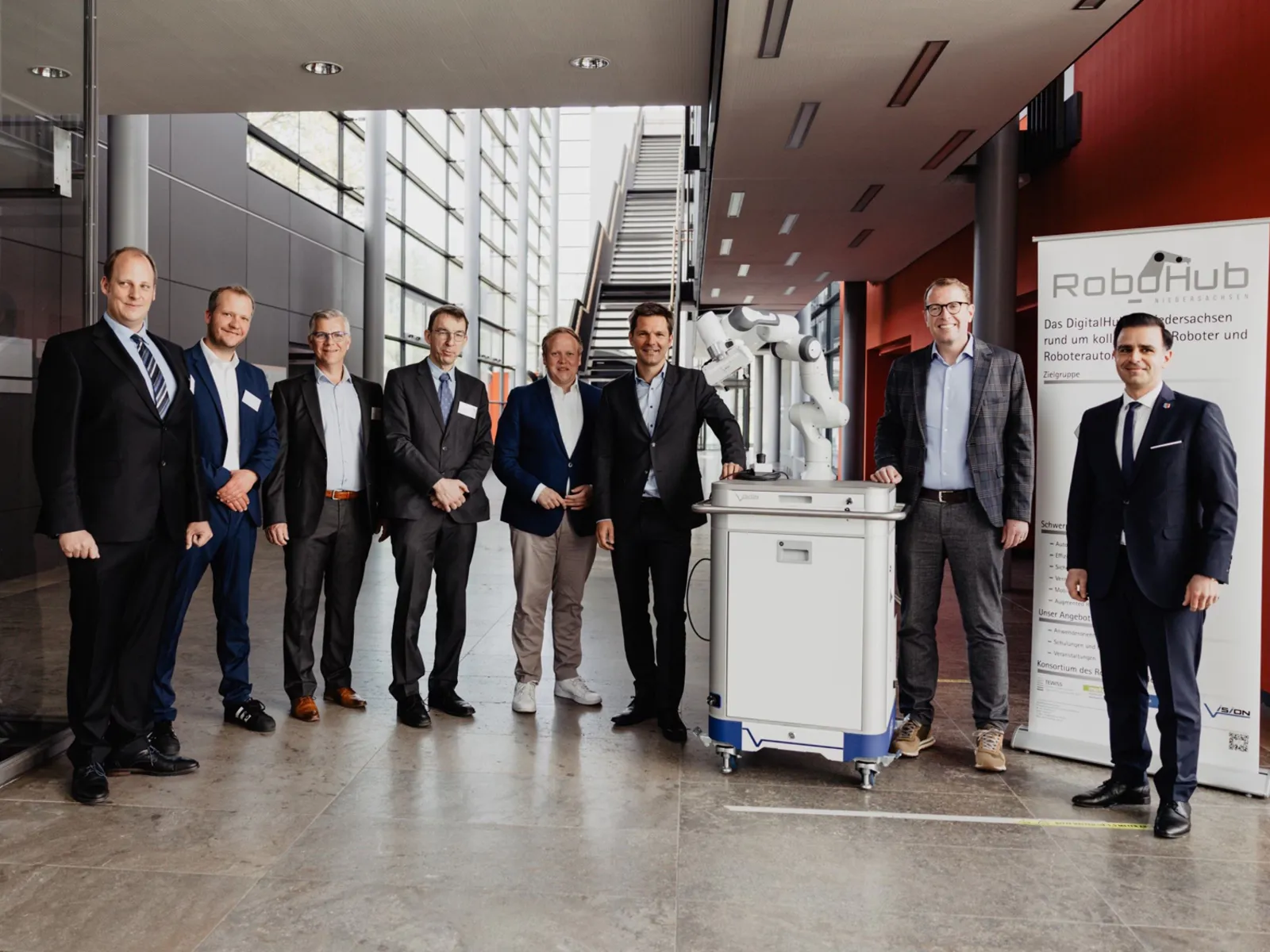 Eight men in suits stand around an industrial robot.