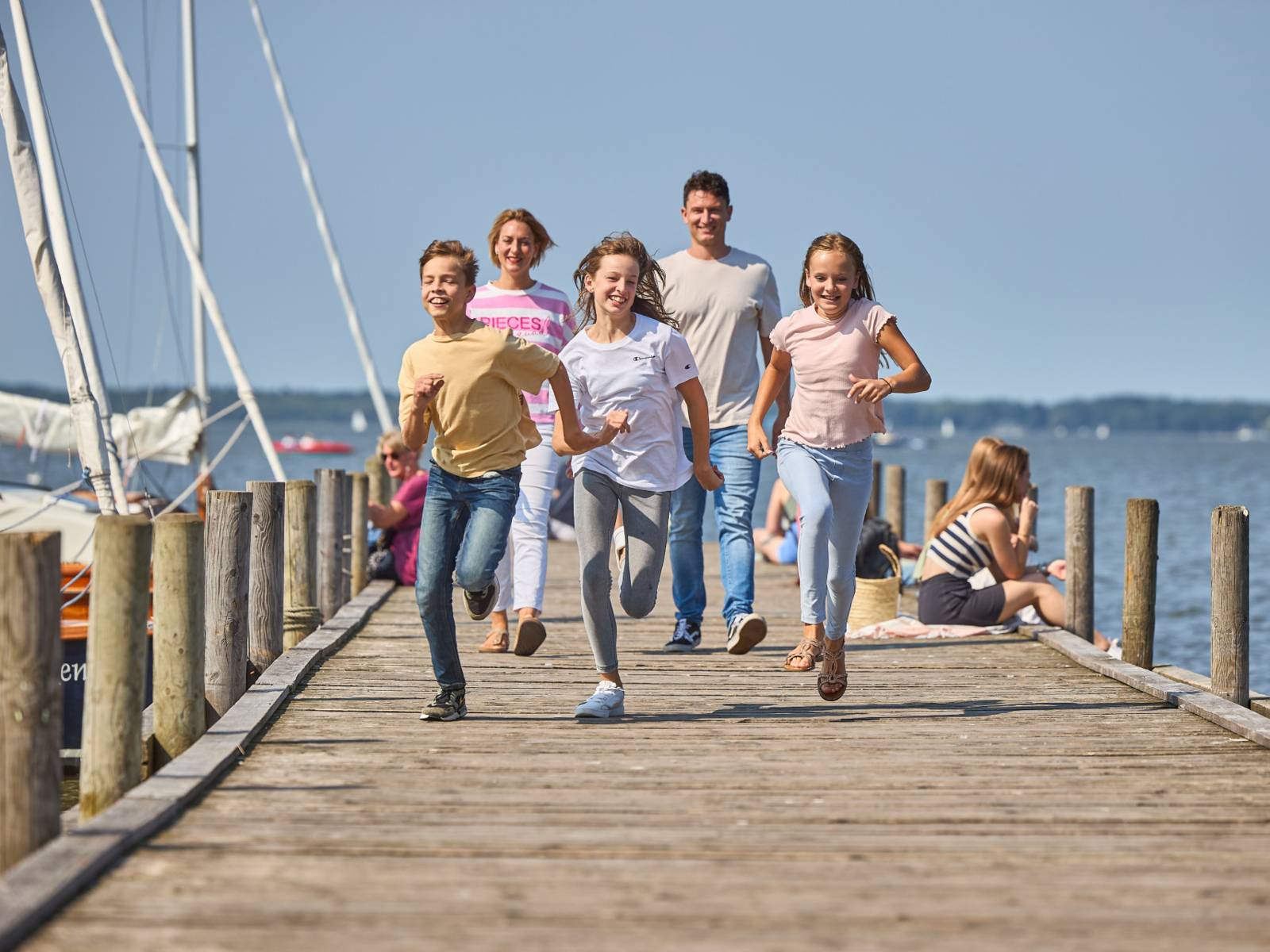 Drei Kinder rennen lachend über einen Steg am See, Eltern folgen im Hintergrund bei sonnigem Wetter.