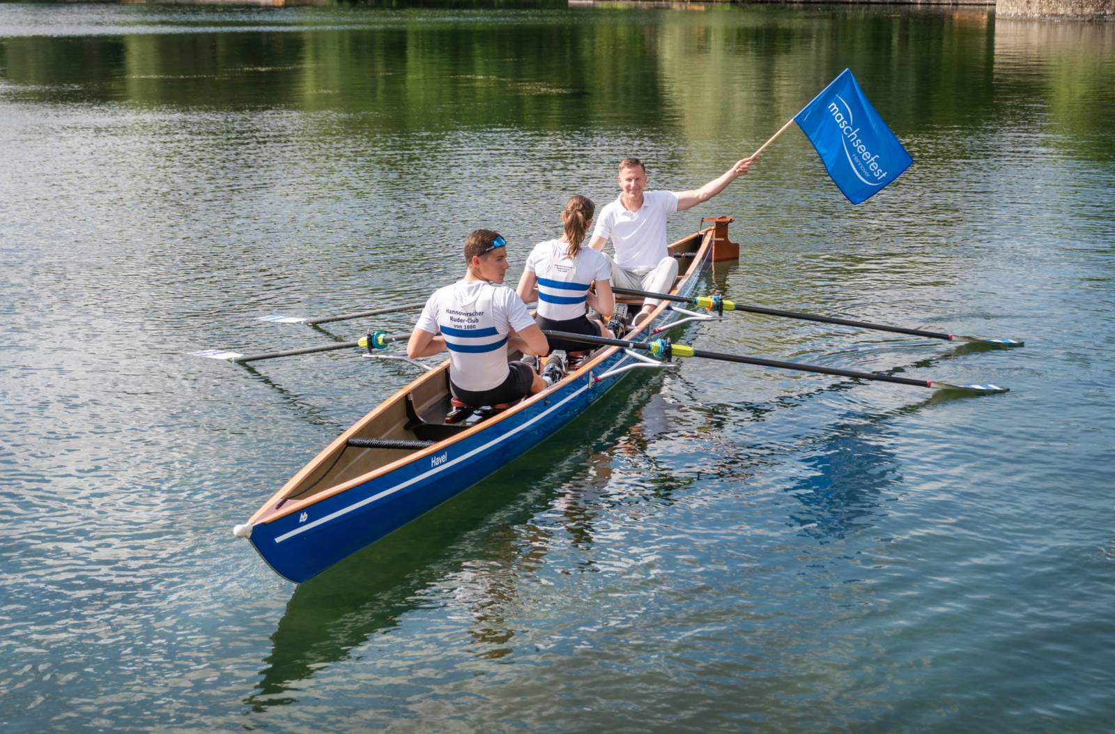 Drei Rudernde und ein Mann mit „Maschseefest“-Fahne in einem Ruderboot auf einem ruhigen See