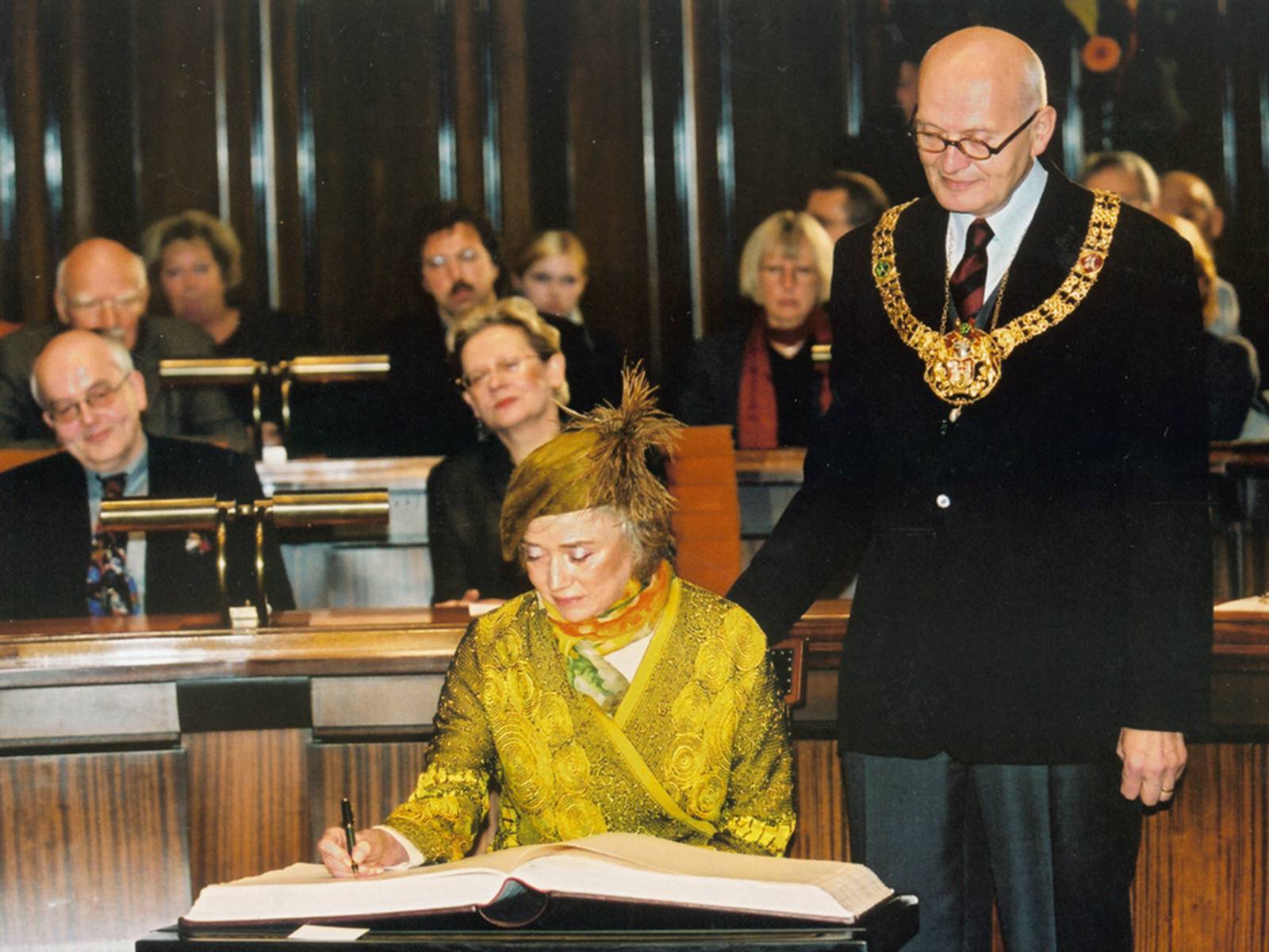 A lady in a yellowish dress signs a book, next to her a bald-headed man watching over her shoulder; many more people in the background