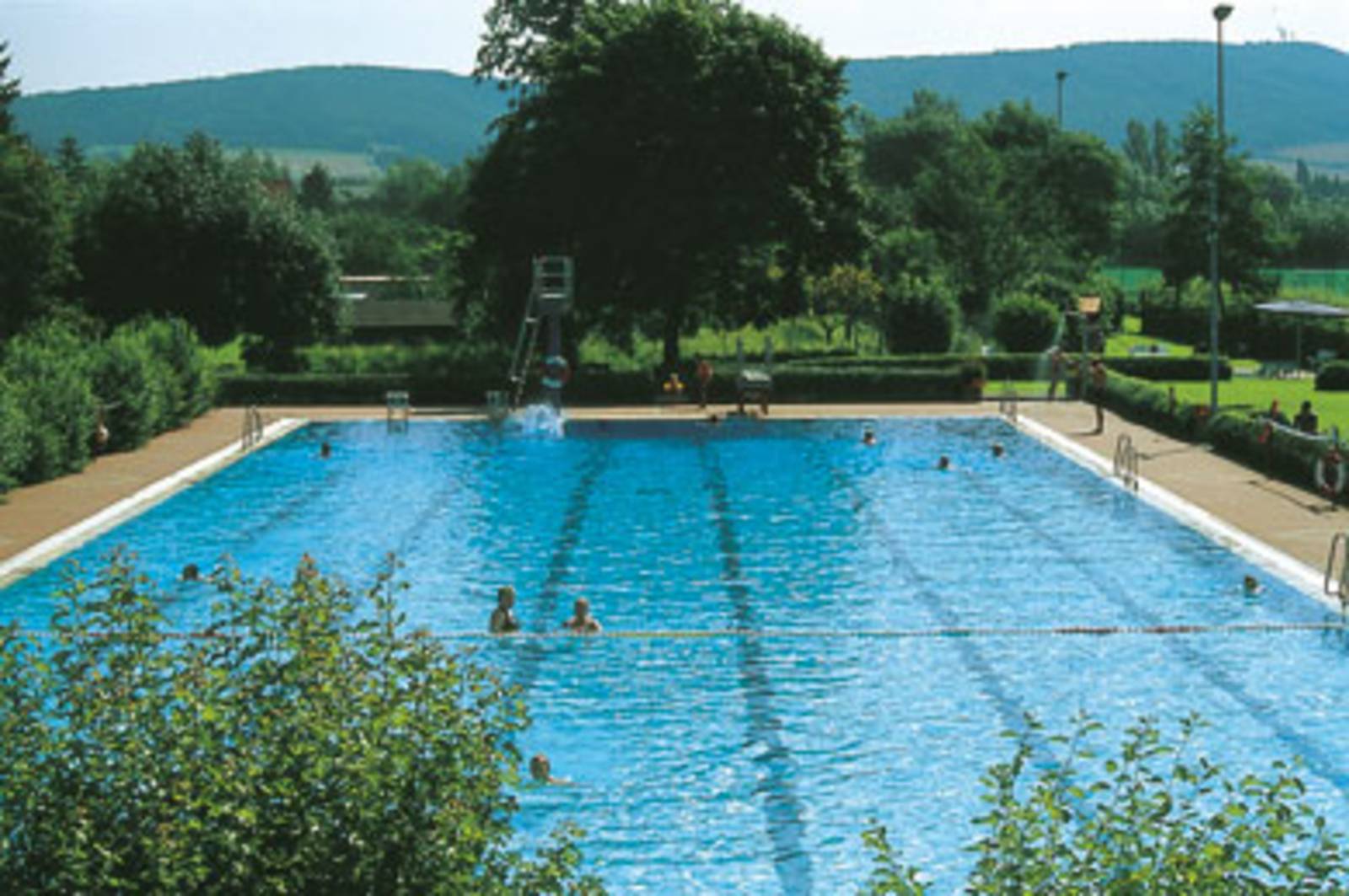 von Bäumen und Rasen umsäumtes Freibad, Berge im Hintergrund 