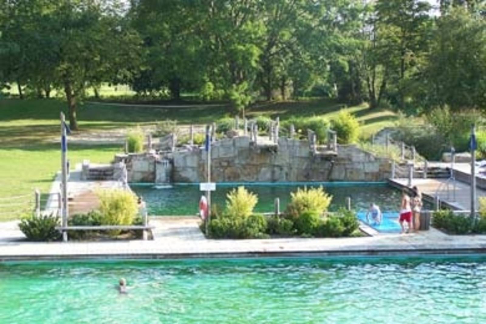 Blick auf eine Holzplattform. Eine Art Steg, der zwei Schwimmbecken voneinander trennt und auf dem Badegäste stehen und der begrünt ist. An das hintere Becken grenzt ein Sprungfelsen mit zwei Sprunghöhen.