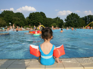 M&auml;dchen mit Schwimmfl&uuml;geln sitzt mit dem R&uuml;cken zum Betrachter am Beckenrand. Im Freibad plantschen viele Kinder.