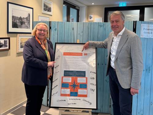 Petra Rosenbach und Hans Nolte präsentieren ein unterschriebenes Nachhaltigkeitsleitbild auf einem Aufsteller im inklusiven Kaffeehaus Anna Leine.