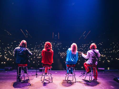 Zwei Männer und zwei Frauen sitzen mit Mikrofonen und Musikinstrumenten auf Barhockern auf einer Bühne.
