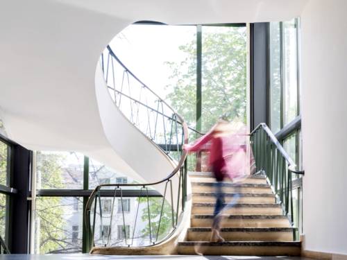 Eine Person steigt eine Treppe hinauf, das Treppenhaus ist hell und in wei&szlig;er Farbe gestrichen, gro&szlig;e Fenster geben den Blick nach drau&szlig;en frei.