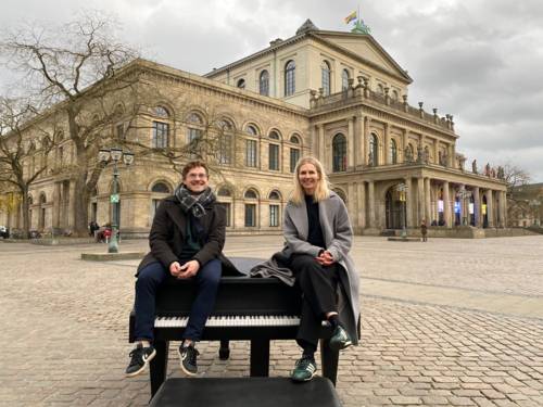 Ein Mann und eine Frau sitzen auf einem schwarzen Klavier, das auf einem weiträumigen Platz steht. Im Hintergrund steht ein historisches Gebäude im spätklassististischen Stil mit Schmuckelementen.