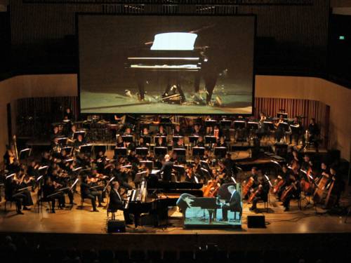 Ein klassisches Orchester auf einer Bühne vor einer Leinwand, auf der das Bild eines zu Boden stürzenden Pianos zu sehen ist.