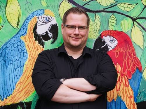 Auf dem Bild ist der Autor Imre Grimm zu sehen. Er steht vor einer Wand die mit Papageien bemalt ist.