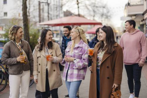 im Vordergrund sind vier Frauen in einer Reihe, die ein Getränk in der Hand halten und sich unterhalten. Im Hintergrund sind weitere personen zu sehen.