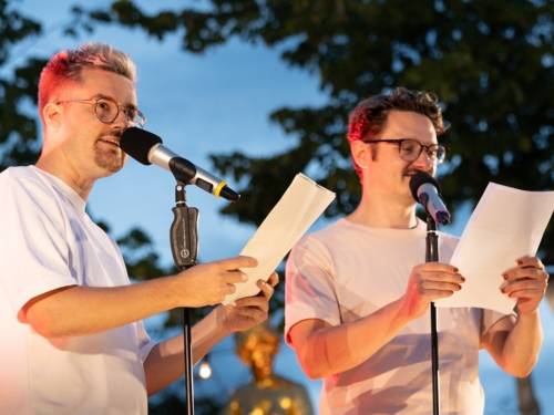 En la imagen aparecen dos participantes en un slam de poesía en un escenario al aire libre, mientras recitan sus textos.