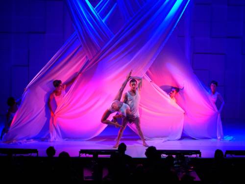 Dancers in white outfits perform in front of artfully draped lengths of fabric, illuminated in blue and pink light.