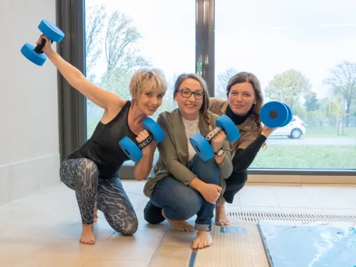 Trois dames, certaines en tenue de sport, sont assises au bord d'une piscine et tiennent des haltères bleus dans leurs mains.