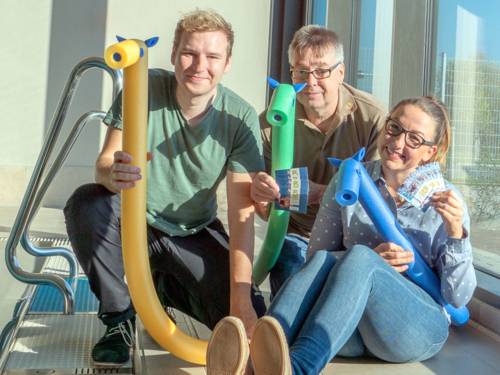 Zwei Männer und eine Frau sind in Straßenkleidung neben einem Schwimmbecken und halten Schwimmnudeln und Postkarten.