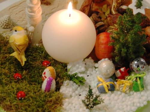 Eine kugelförmige Kerze brennt inmitten verschiedener Dekorationsartikel, darunter Vögel aus Stoff, Marienkäfer und Eier aus Holz, Moos und Geschenkepäckchen.