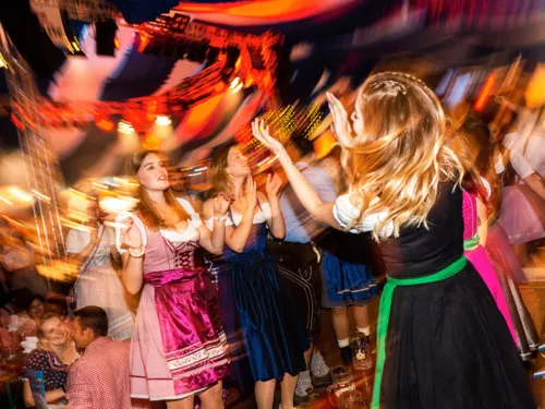 Ausgelassene Stimmung auf einem Oktoberfest mit tanzenden Frauen in Dirndln, bunten Lichtern und verschwommenen Bewegungen.