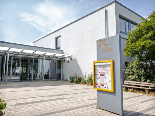 Im gepflasterten Eingangsbereich zur Brüder-Grimm-Schule steht eine Schautafel mit Aktivitäten der Schule. Die Teilansicht der Schule zeigt ein weiß verputztes, rechteckiges, zweistöckiges Gebäude mit großen Fenstern.