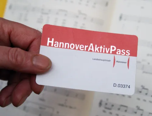 Em primeiro plano, vê-se uma mão a segurar o «Hannover-Aktiv-Pass» vermelho e branco; ao fundo, encontram-se partituras