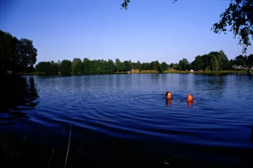 Ansicht des Sees, in dem zwei Menschen schwimmen. Im Hintergrund befinden sich viele Bäume.