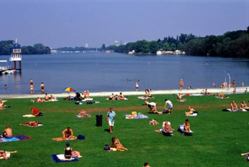im Vordergrund liegen und sitzen Menschen in Badebekleidung auf Badetüchern auf Rasen, im Hintergrund ist ein dünner Streifen Strand und der Maschsee