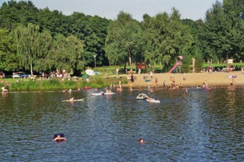 Im See schwimmende und spielende Personen, im Strandbereich Badegäste und Spielgeräte, dahinter Liegewiese und Wald