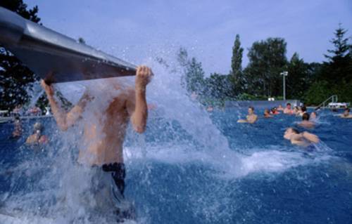 Ein Junge hat sich am Kopf der Wasserschwallbrause hochgezogen, der Körper wird vom Wasser umströmt, im Hintergrund im Wasser spielende Kinder