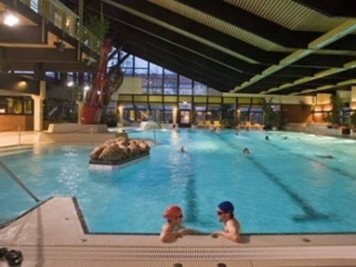 Innenansicht des Hallenbades mit Blick auf das Schwimmerbecken. Es sind wenige Menschen im Wasser. Eine Frau lehnt sich an eine kleine Insel im Vordergrund.