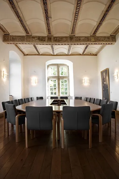 Ein lichtdurchfluteter Saal in einem Schloss mit einem gro&szlig;en Besprechungstisch, andem zwanzig St&uuml;hle stehen.