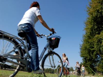 Eine Radfaherin fährt durch Felder und Wiesen, ihr kommen junge Radfahrerinnen entgegen.