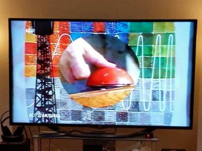 Fernsehbild in Farbe, das einen einen Daumen, der auf einen Knopf drückt, zeigt