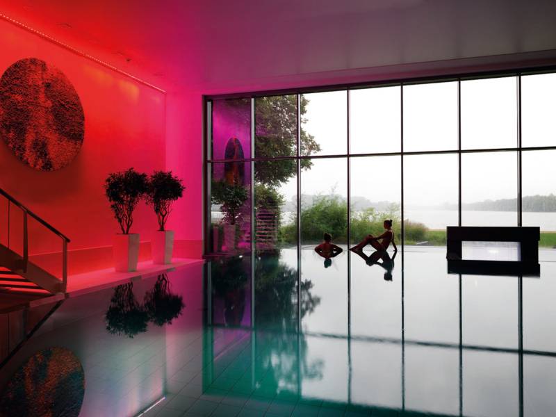 Blick über die Wasserfläche eines Schwimmbeckens, am hinteren Beckenrand sind zwei junge Frauen, dahinter ist eine große Fensterfläche. Durch die Fenster ist der Maschsee zu sehen.