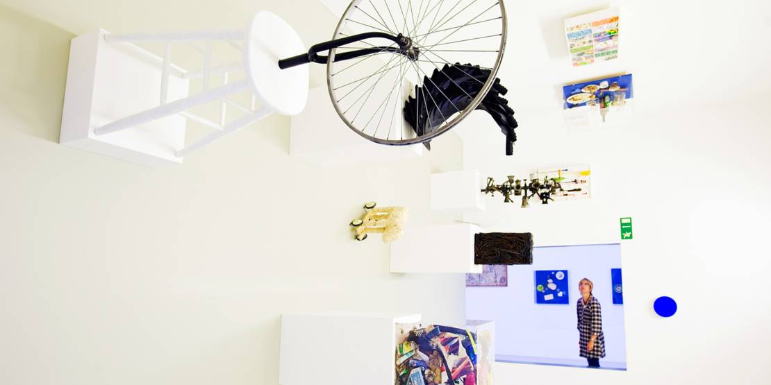 A woman looks through an opening into a room in which art objects are mounted on a wall, creating the impression that the room has been rotated by 90 degrees.