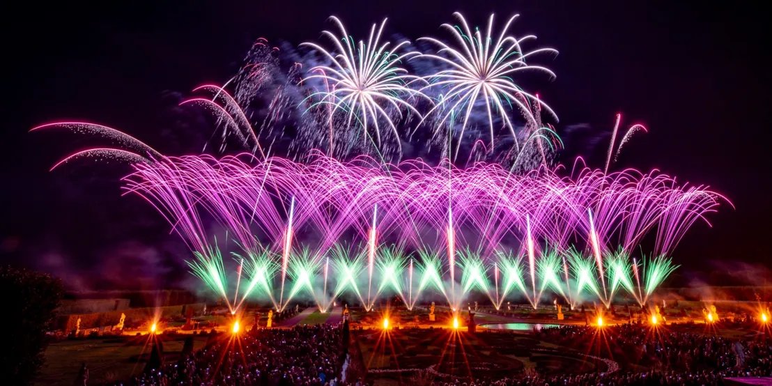 The picture shows a firework display in the Herrenhausen Gardens, performed in bright colors of pink and green in front of a large audience. The spectators can be seen in the foreground, while the fireworks illuminate the sky above the historic gardens in the background.