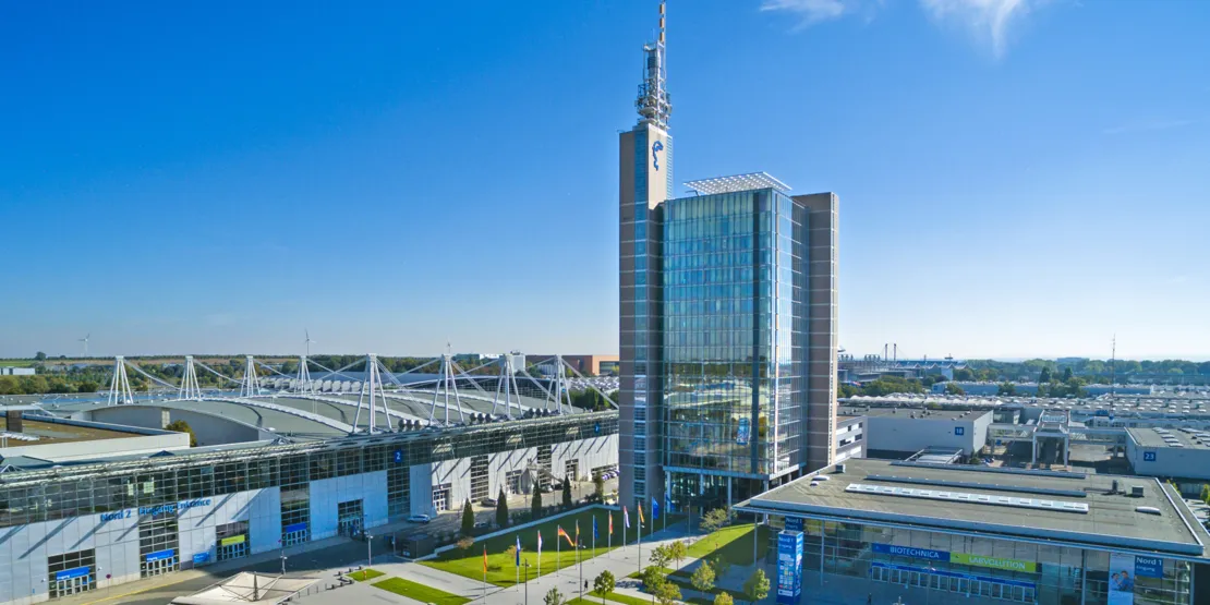 Hannover Fuar Alanı'nın havadan çekilmiş görüntüsü; modern cam bina, radyo kulesi ve dikkat çekici çatı yapılarına sahip salonlar.