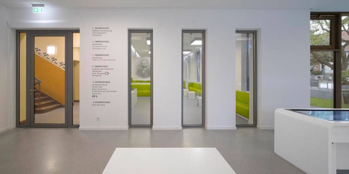 Ein Foyer mit einem Medientisch. Rechts neben der Tür, die in ein Treppenhaus und zu weiteren Räumlichkeiten führt, ist an die Wand geschrieben, was in den einzelnen Stockwerken zu finden ist.