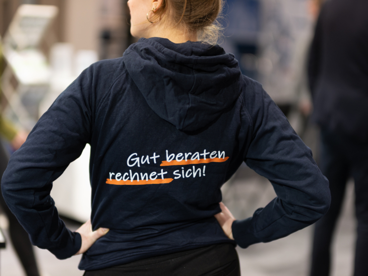 Une jeune femme, de dos face à la caméra, porte un sweat à capuche bleu sur lequel on peut lire : « Bien se faire conseiller, ça vaut le coup ! »