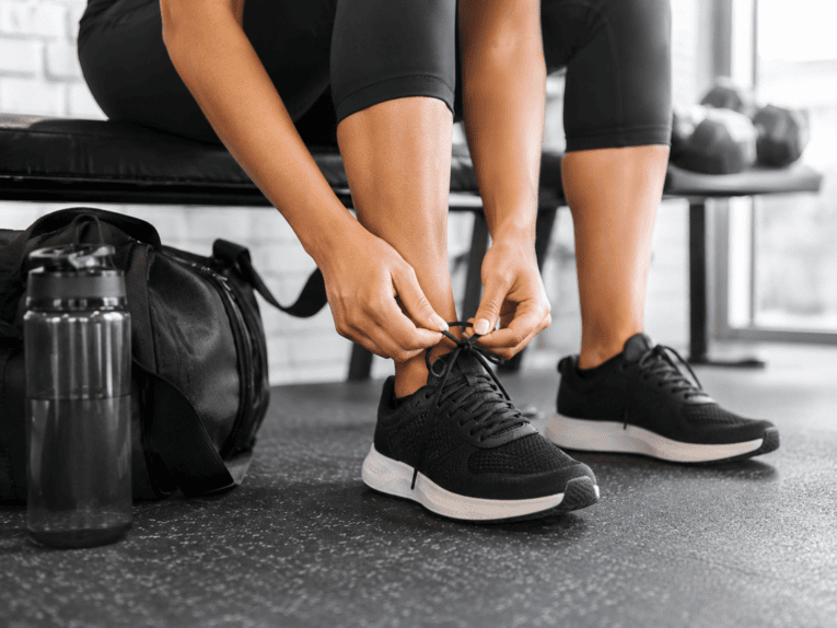 Una persona è seduta su una panchina da palestra nera. Si vedono solo le gambe. La persona si sta allacciando le scarpe da ginnastica nere. Accanto a lei ci sono una bottiglia d’acqua e una borsa da palestra.