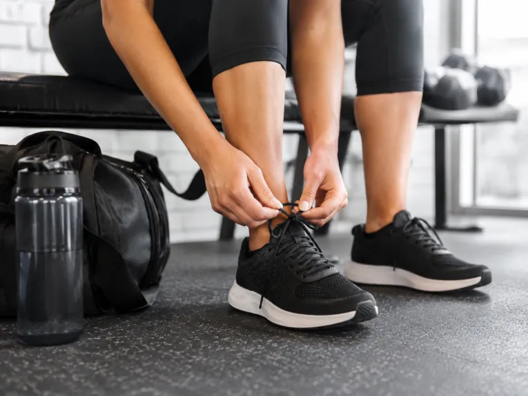 Una persona è seduta su una panchina da palestra nera. Si vedono solo le gambe. La persona si sta allacciando le scarpe da ginnastica nere. Accanto a lei ci sono una bottiglia d’acqua e una borsa da palestra.