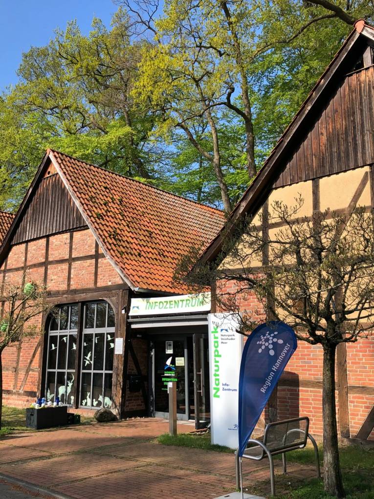 Mehrere historische Fachwerkscheunen stehen nebeneinander. Zwischen zwei Scheunen ist eine Glastür, darüber ein Banner mit dem Text: "INFOZENTRUM". In der Nähe der Eingangstür sind noch weitere Aufsteller, Displays etc. mit dem Logo des Naturparks Steinhuder Meer oder der Region Hannover.