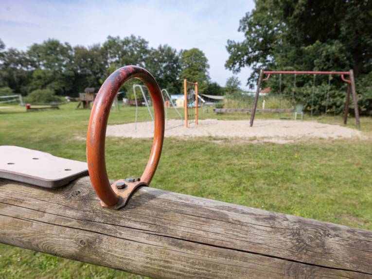 Im Vordergrund Griff und Holzbalken einer Wippe, im Hintergrund Schaukeln, Klettermöglichkeiten, Fussballtor