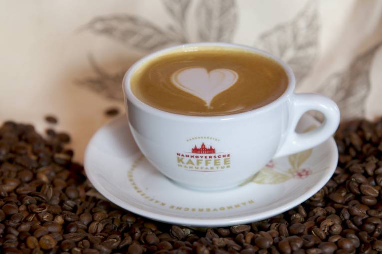 Hannoversche Kaffeemanufakturi kujundusega tass on täis kohvi ja sellel on valgel laual alustassil südamekujuline latte art. Alustassi ääristavad kohviubad.
