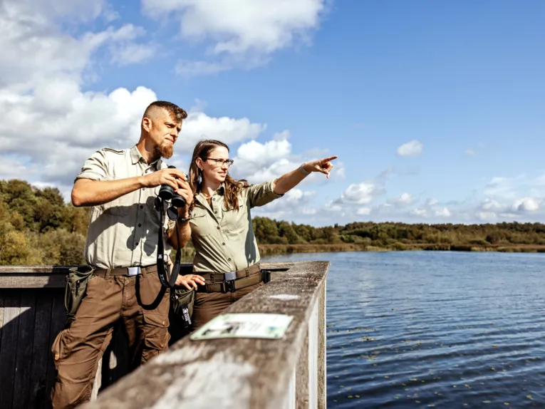 Un ranger e una donna sono in piedi su una piattaforma panoramica. Lui tiene in mano un binocolo, lei punta in una direzione.