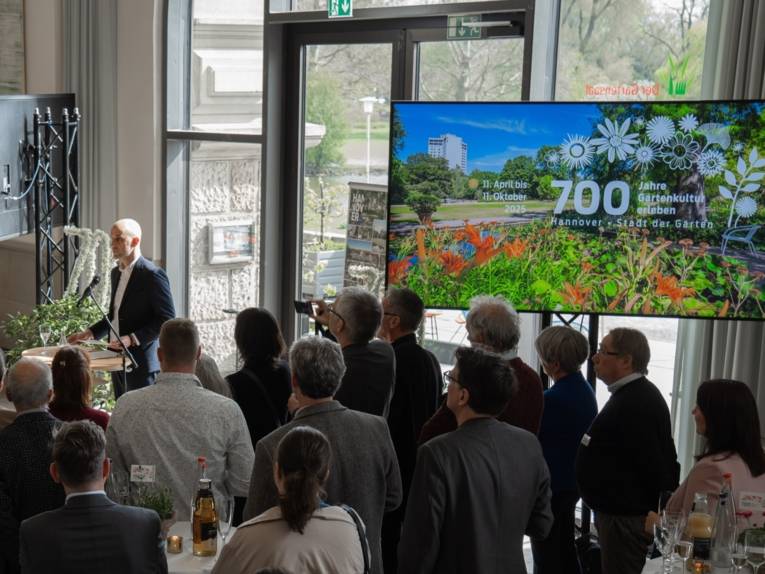 Eine Menge Menschen steht in einem Halbkreis um eine Person, die eine Rede hält. Auf einem Bildschirm am rechten Bildrand steht "700 Jahre Gartenkultur erleben; Hannover - Stadt der Gärten"