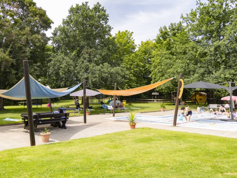 Mit bunten Tüchern überdachter Bereich, in dem sich ein großes Planschbecken befindet. Drumherum Rasenfläche mit Baumbestand.