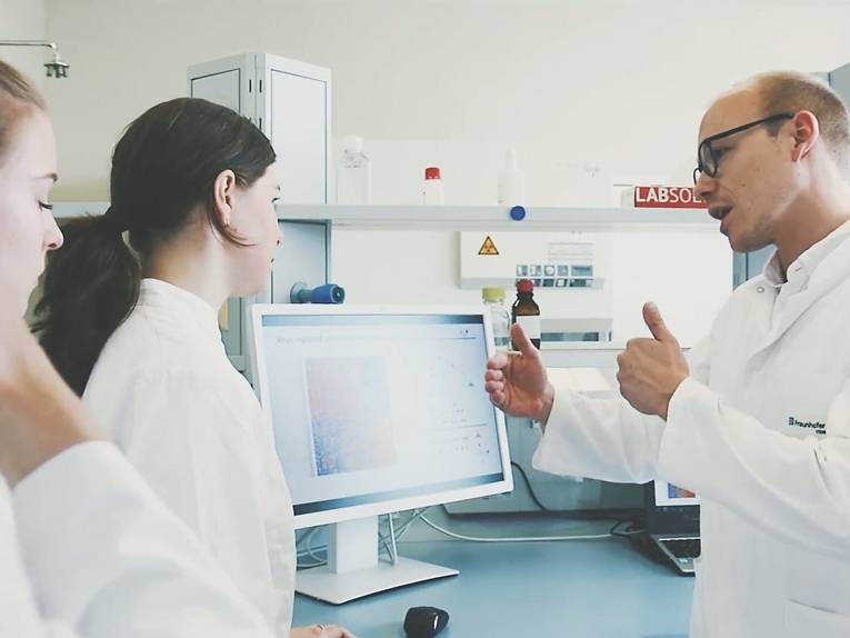 Ein Mann referiert für mehreren Personen vor einem Computerbildschirm. Alle Personen tragen Laborkleidung.